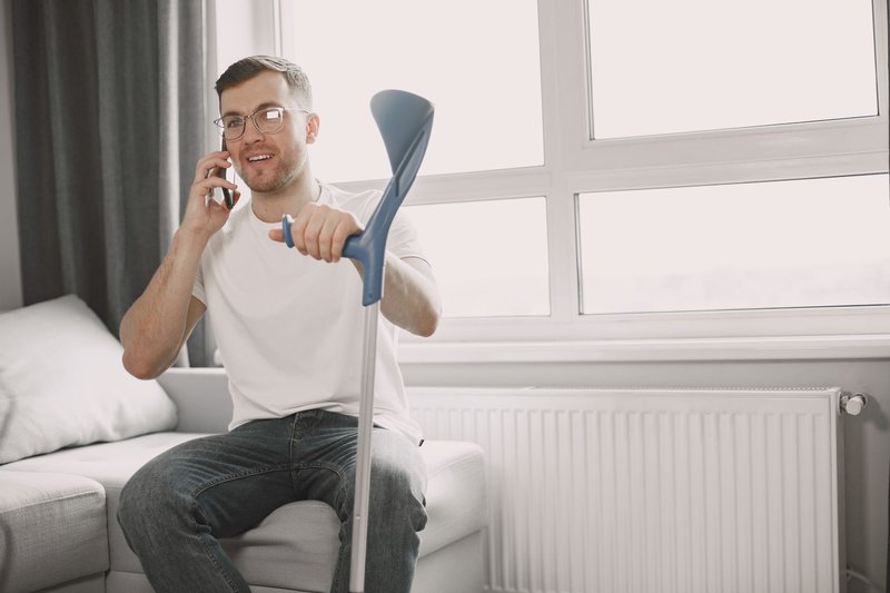 Mann mit Gehstütze sitzt zu Hause auf dem Sofa und telefoniert