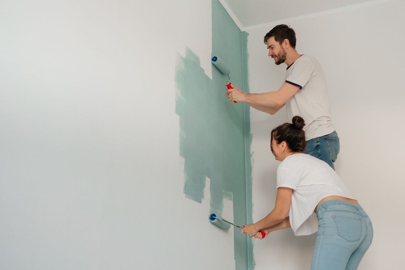 Ein Mann und eine Frau streichen mit Farbrollen gemeinsam eine Zimmerwand grün