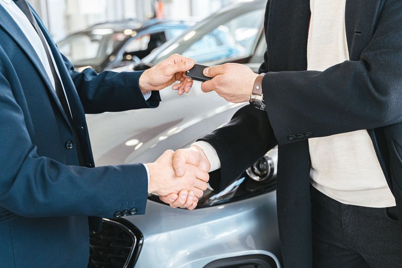 Zwei Personen geben sich vor einem silbernen Auto die Hand, dabei wechselt ein Autoschlüssel den Besitzer