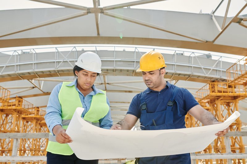Zwei Personen mit Schutzhelmen studieren auf einer Baustelle einen großen Bauplan