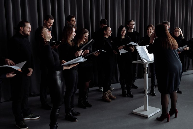 Ein Chor aus Erwachsenen in dunkler Kleidung singt mit Notenmappen vor einer Dirigentin am Notenpult