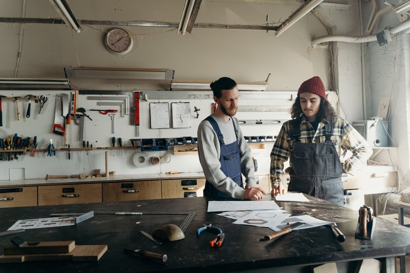 Zwei Handwerkerinnen und Handwerker in Latzhosen stehen an einer Werkbank und besprechen eine Zeichnung