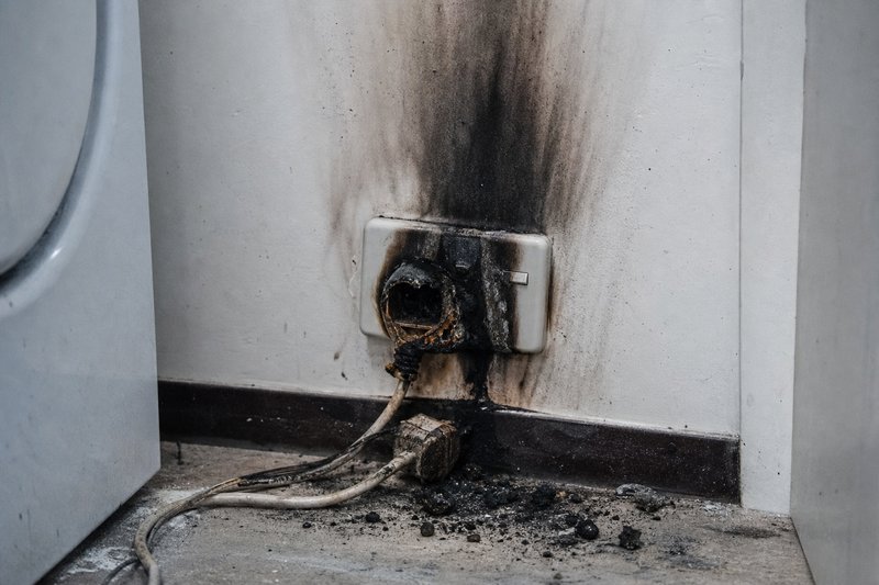 Ausgebrannte Steckdose mit Rußspuren an der Wand über dem verschmorten Stecker neben einer Waschmaschine