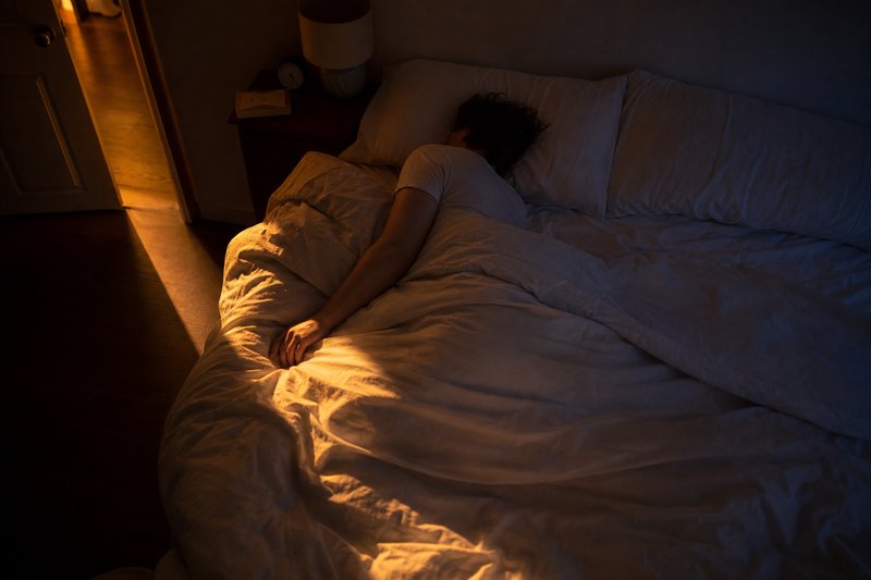 Schlafende Person im Bett, ein schmaler Lichtstreifen aus der halb offenen Zimmertür fällt auf die Bettdecke