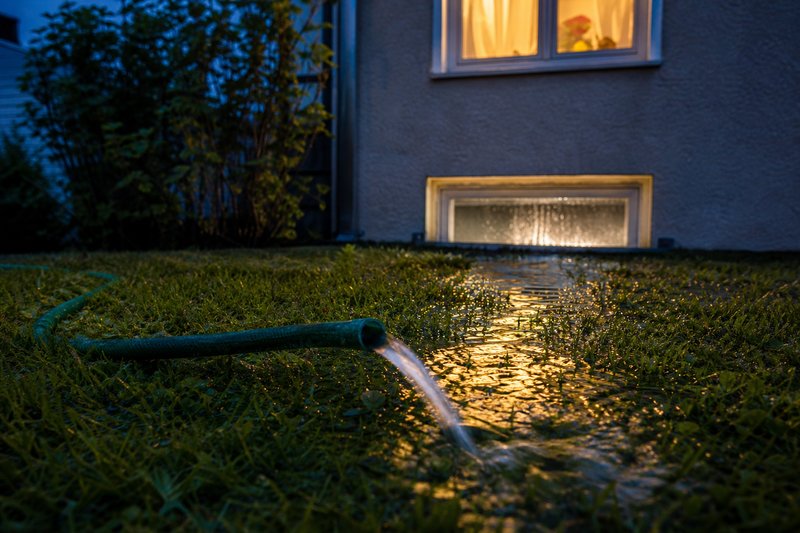 Aus einem Gartenschlauch läuft Wasser über den Rasen auf ein beleuchtetes Kellerfenster zu