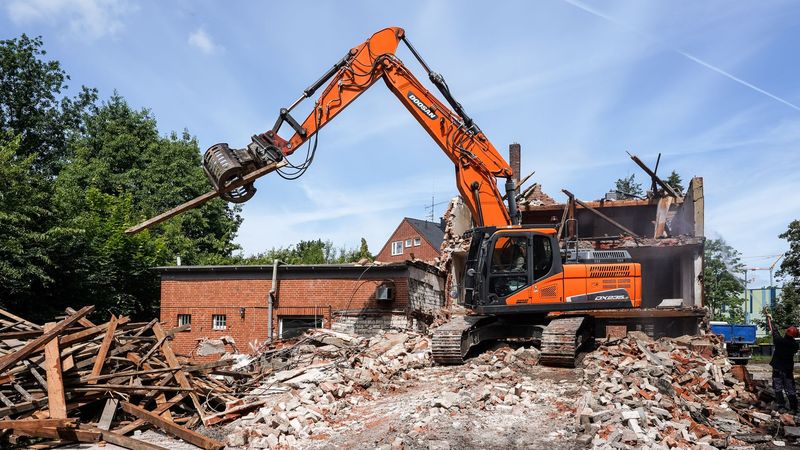 Orangefarbener Abrissbagger reißt ein Backsteinhaus ein, davor ein Haufen aus Ziegeln und Balken