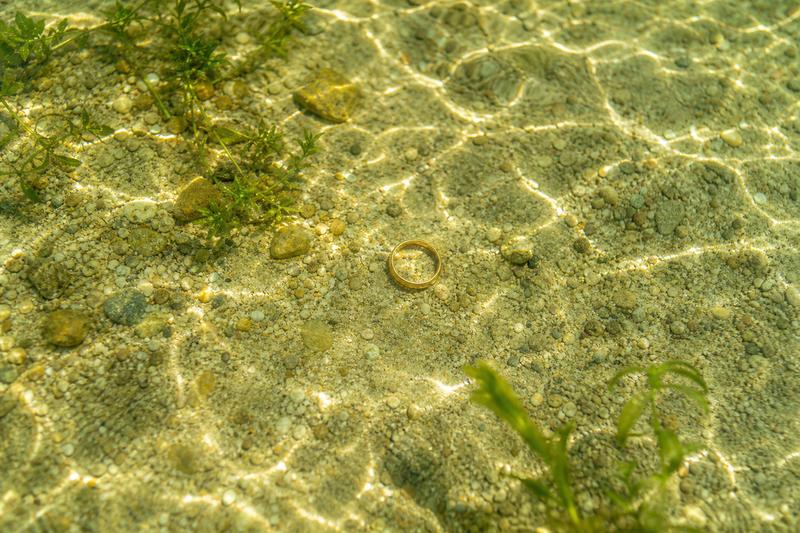 Goldener Ehering auf dem sandigen Grund eines flachen Sees
