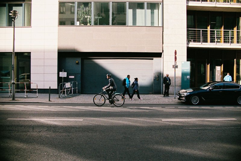 Radfahrer ohne Helm fährt auf einer Berliner Straße an parkenden Autos und Fußgängern vorbei