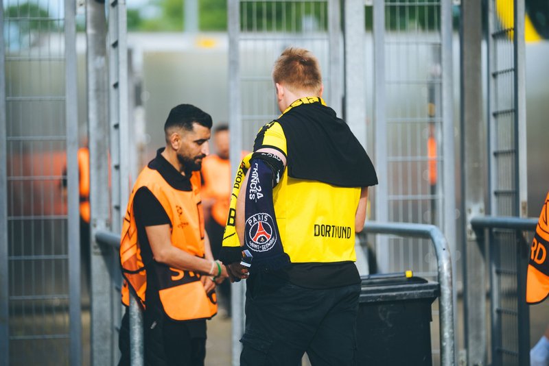 Ordner in orangen Warnwesten kontrollieren den Einlass zu einem Fußballstadion, ein Fan im Vereinstrikot passiert die Sperre