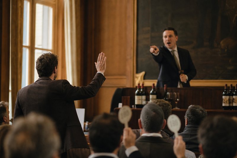 Mann hebt in einem Auktionssaal die Hand, während der Auktionator vom Podium aus auf ihn zeigt, im Vordergrund Bieter mit Nummernschildern