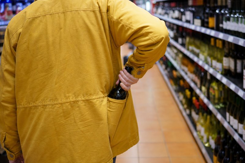 Person in gelber Jacke von hinten schiebt im Getränkegang eines Supermarkts eine Weinflasche in die Jackentasche