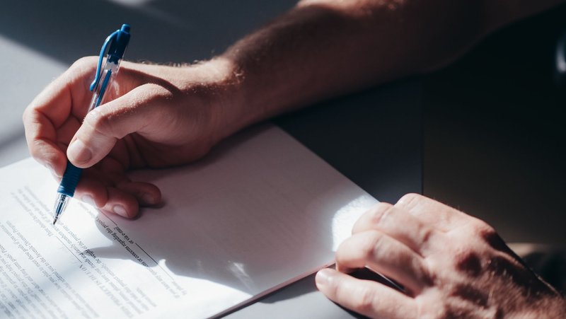 Nahaufnahme einer Hand, die mit blauem Kugelschreiber ein Schriftstück auf dem Tisch unterschreibt