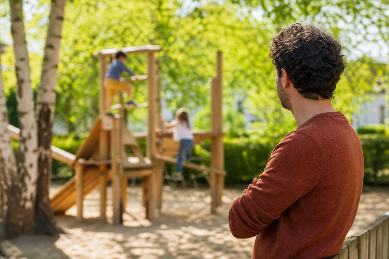 Vater am Rand eines Spielplatzes beobachtet aufmerksam seine beiden Kinder auf einem Klettergerüst