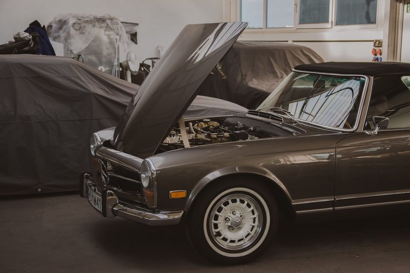 Klassischer Mercedes mit geöffneter Motorhaube in einer Garage, im Hintergrund abgedeckte Fahrzeuge
