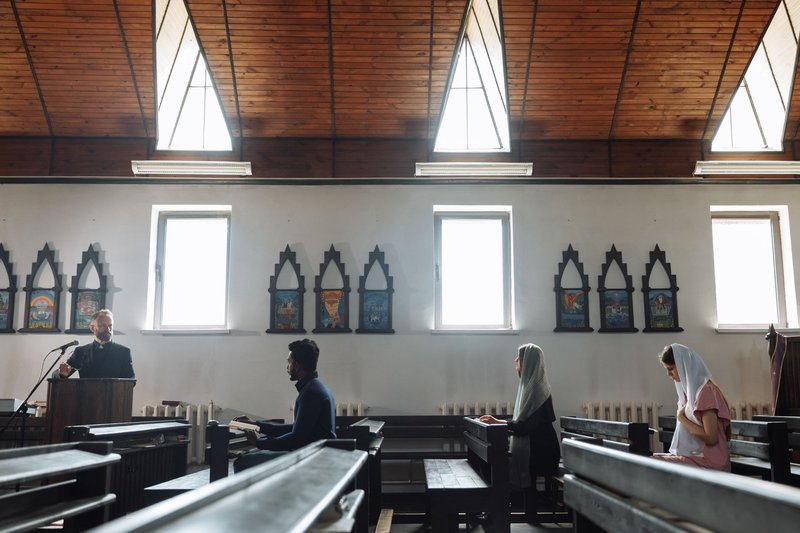 Schlichter Kirchenraum, ein Geistlicher am Ambo, einzelne Besucher sitzen in den Bänken und beten