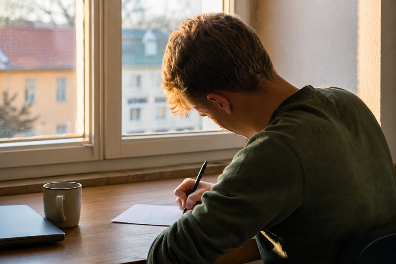 Junger Mann sitzt am Schreibtisch vor dem Fenster und schreibt von Hand einen längeren Text auf liniertes Papier