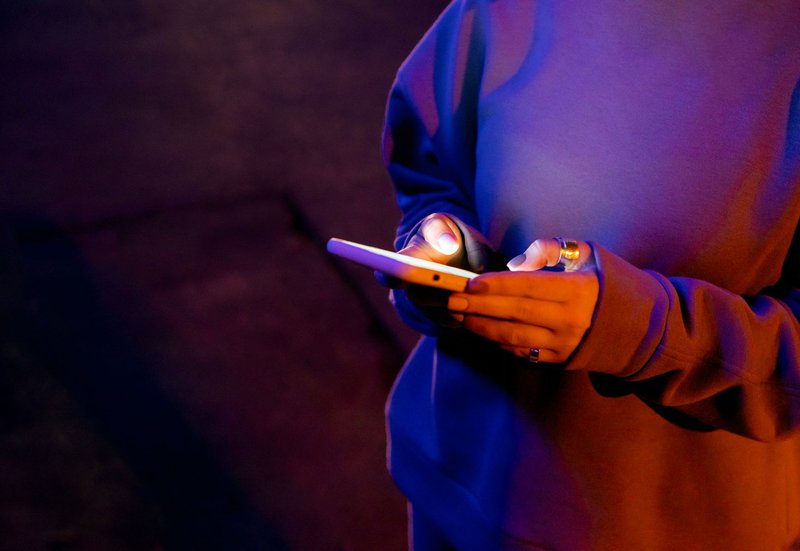 Hände tippen im Dunkeln eine Nachricht auf einem Smartphone, der Bildschirm leuchtet in Violett und Orange