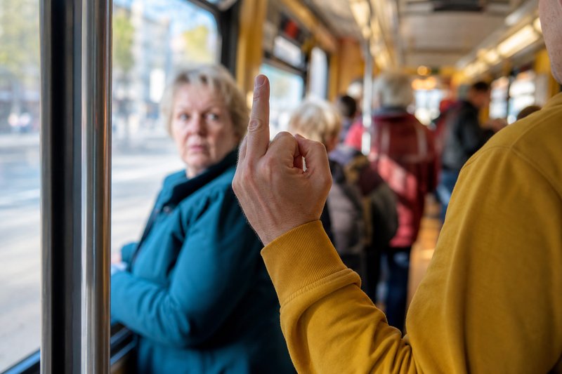 Erhobene Hand mit ausgestrecktem Mittelfinger in einer Straßenbahn, im Hintergrund blickt eine Frau in Richtung der Geste