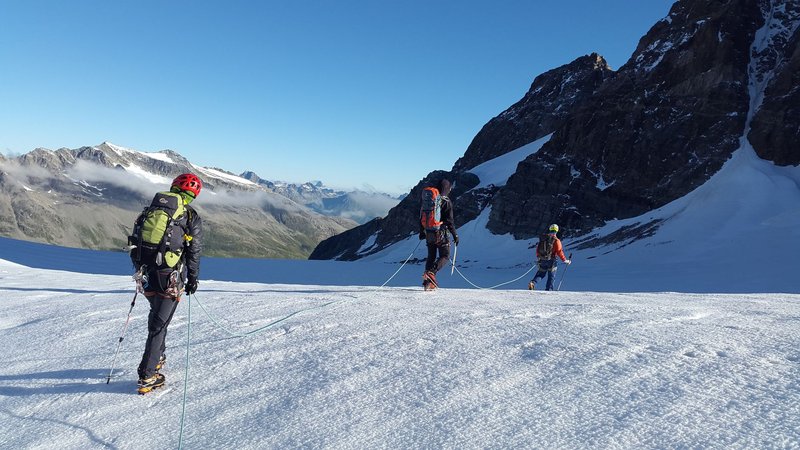 Drei angeseilte Bergsteiger gehen hintereinander über ein Gletscherfeld vor Felswänden