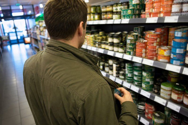 Mann von hinten in einem Supermarktgang, der eine kleine Dose in seine Jackentasche steckt