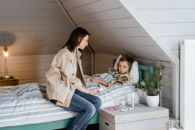 Mutter sitzt am Bett ihres kranken Kindes, daneben Fieberthermometer und Medikamente