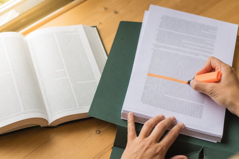 Hände markieren mit einem orangen Textmarker eine Passage in einem ausgedruckten Urteil neben einem aufgeschlagenen Gesetzbuch