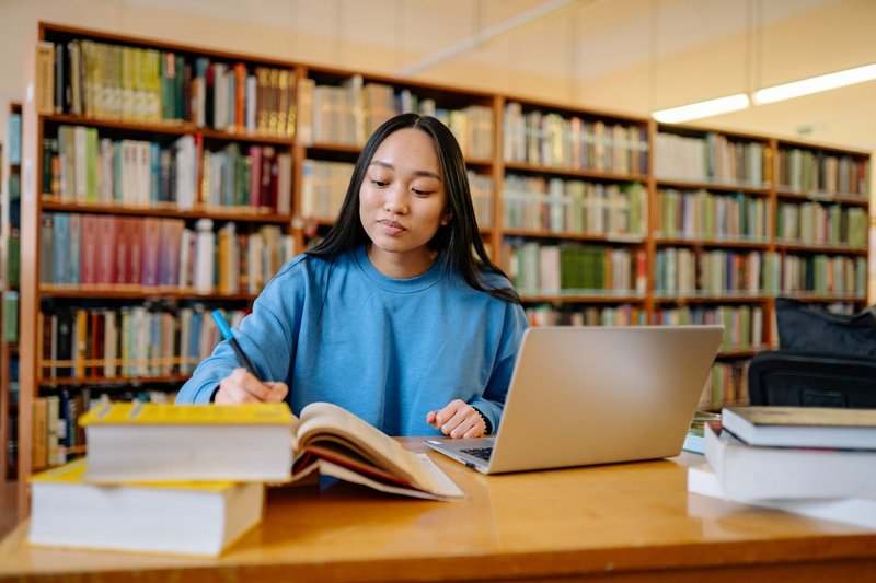 Studentin sitzt in einer Bibliothek am Laptop und blättert dabei in einem aufgeschlagenen Buch