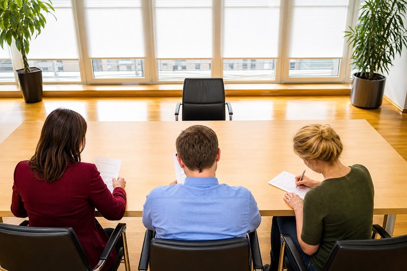 Dreiköpfige Auswahlkommission von hinten an einem Konferenztisch, gegenüber steht ein leerer Stuhl