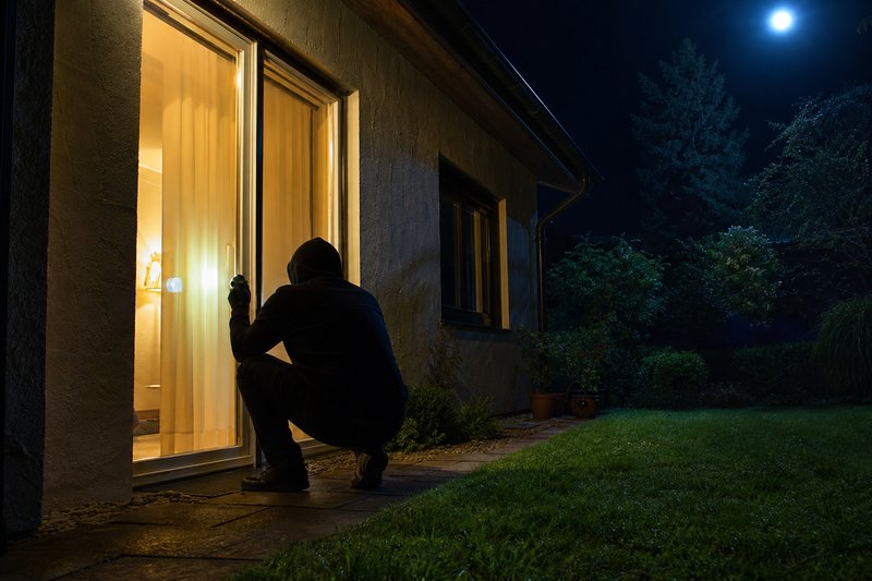 Dunkle Gestalt kauert nachts vor der beleuchteten Terrassentür eines Einfamilienhauses und leuchtet mit einer Taschenlampe an die Scheibe