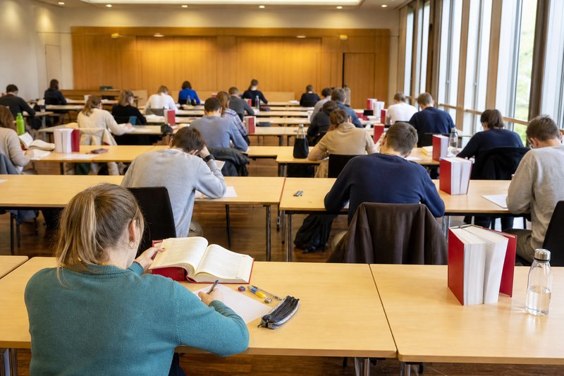 Studierende schreiben in einem Klausursaal mit langen Tischreihen, im Vordergrund blättert eine Studentin im aufgeschlagenen Gesetzestext