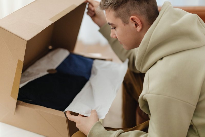 Junger Mann öffnet zu Hause einen Karton und zieht ein Kleidungsstück heraus