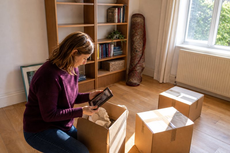 Frau kniet in einer geräumten Wohnung vor einem Umzugskarton und hält ein gerahmtes Foto in den Händen