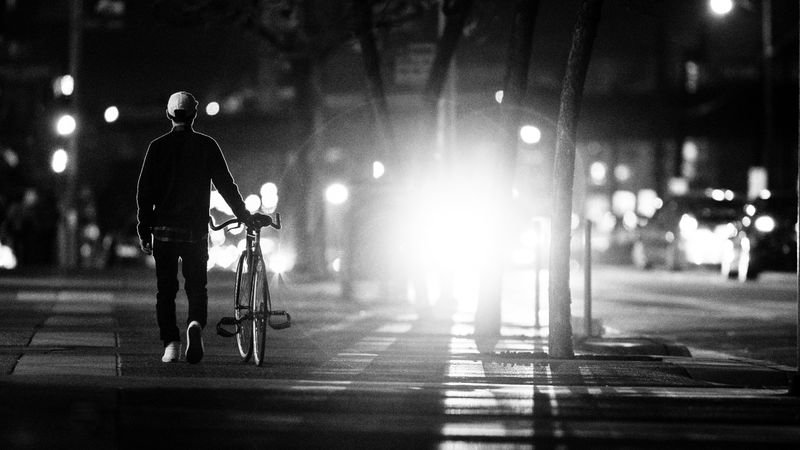 Person schiebt nachts ein Fahrrad über den Gehweg einer beleuchteten Stadtstraße