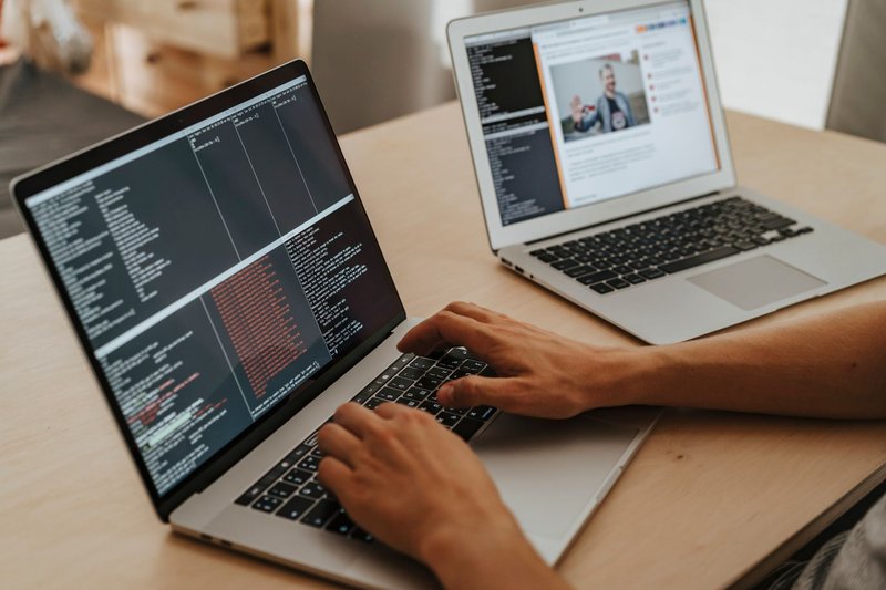 Hände tippen an einem Laptop mit Programmcode, daneben ein zweiter aufgeklappter Laptop