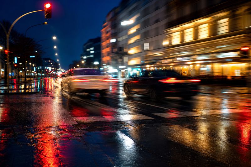 Zwei Autos beschleunigen nebeneinander über eine nasse Stadtkreuzung bei Nacht, Lichter zu Streifen verwischt