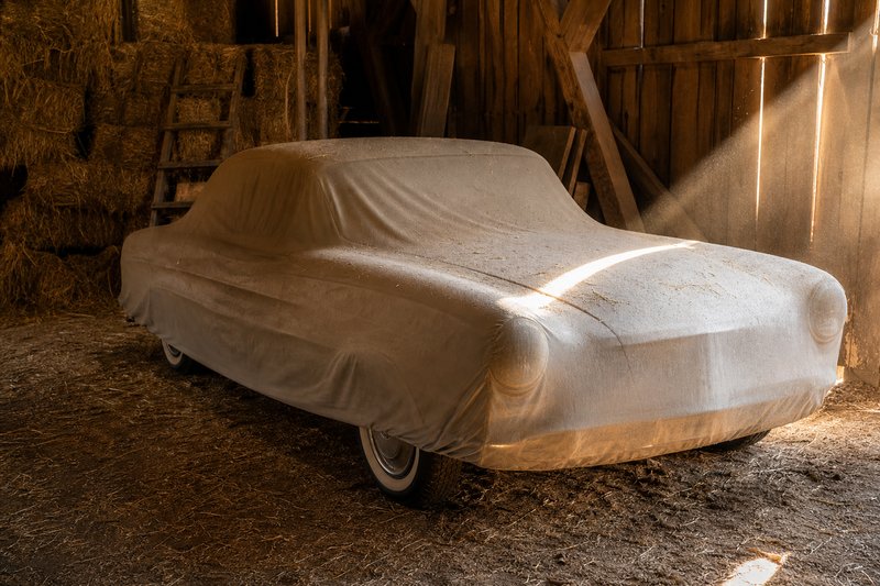 Mit einer hellen Plane abgedeckter Oldtimer in einer Scheune, Sonnenlicht fällt durch die Bretterwand