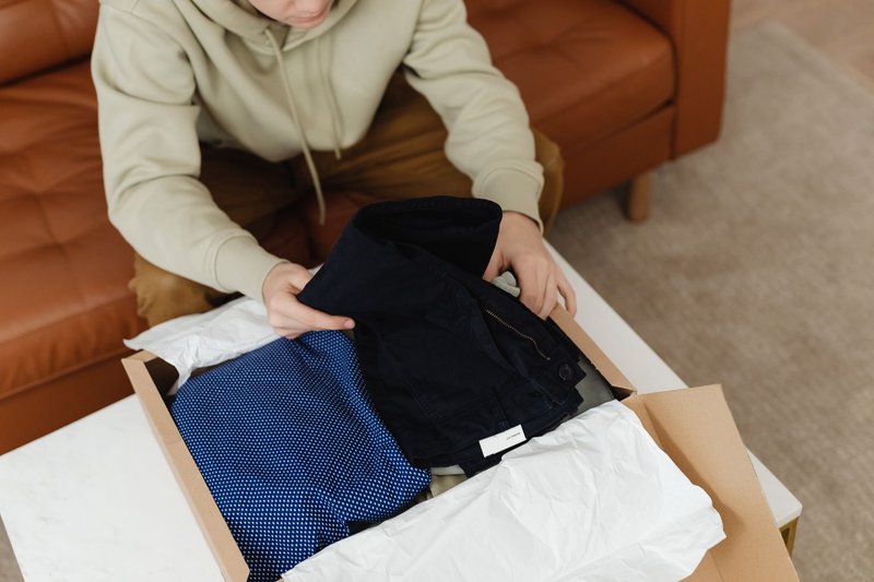 Person sitzt zu Hause auf dem Sofa und nimmt ein dunkles Kleidungsstück aus einem geöffneten Versandkarton