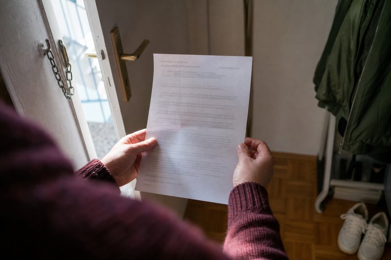 Hände halten in einem Wohnungsflur ein amtliches Schreiben, im Hintergrund die halb geöffnete Wohnungstür mit Sicherheitskette