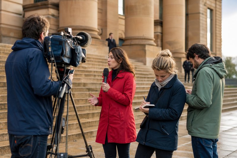 Kamerateam und zwei Journalistinnen berichten auf der Freitreppe eines Gerichtsgebäudes, eine Reporterin im roten Mantel spricht ins Mikrofon