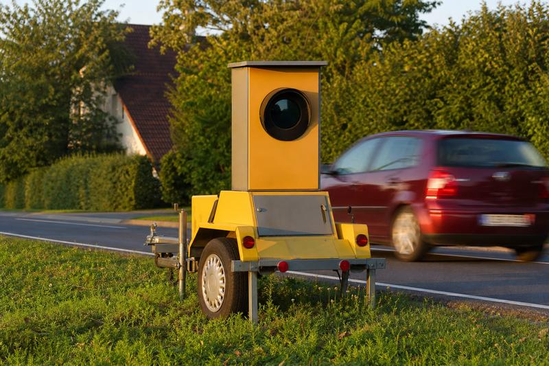Gelber Blitzeranhänger am Grasstreifen einer Ortsdurchfahrt, ein rotes Auto fährt in der Abendsonne unscharf vorbei