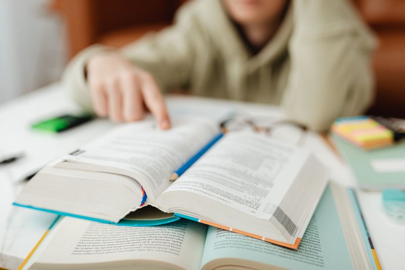 Studierender zeigt mit dem Finger auf eine Stelle in einem aufgeschlagenen dicken Buch, daneben Notizen, Klebezettel und eine Brille