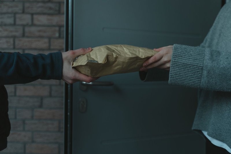An einer dunklen Wohnungstür reicht eine Person eine Sendung in die Hände einer anderen Person