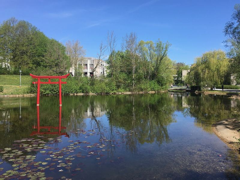 Rotes Torii im Uni-See der Universität Augsburg, dahinter Campusgebäude und Bäume