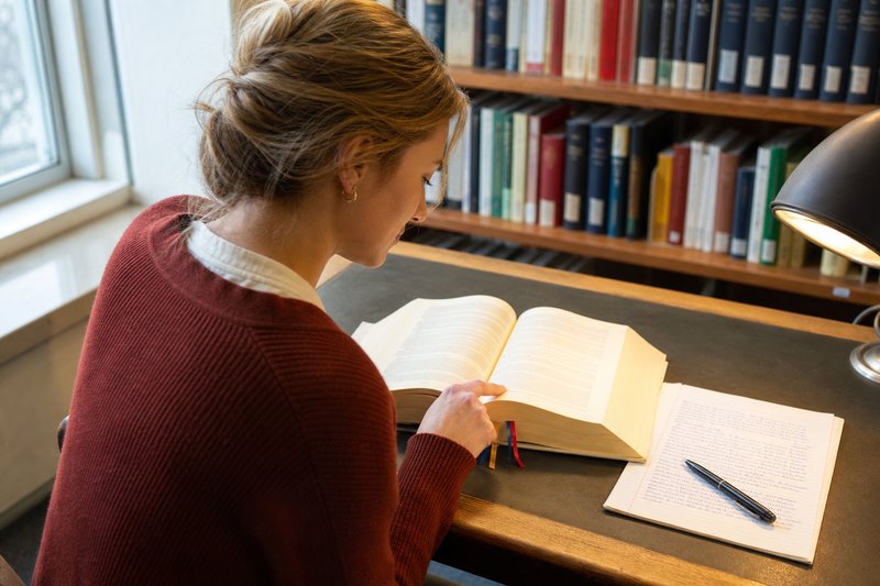 Studentin in rotem Cardigan blättert am Bibliothekstisch in einer aufgeschlagenen Gesetzestextausgabe, daneben ein handbeschriebener Bogen