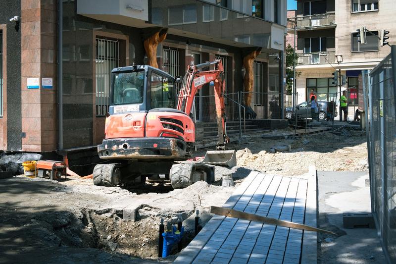 Offene Baugrube mit Leitungsrohr in einer Stadtstraße, daneben ein Bagger und Wohnhäuser