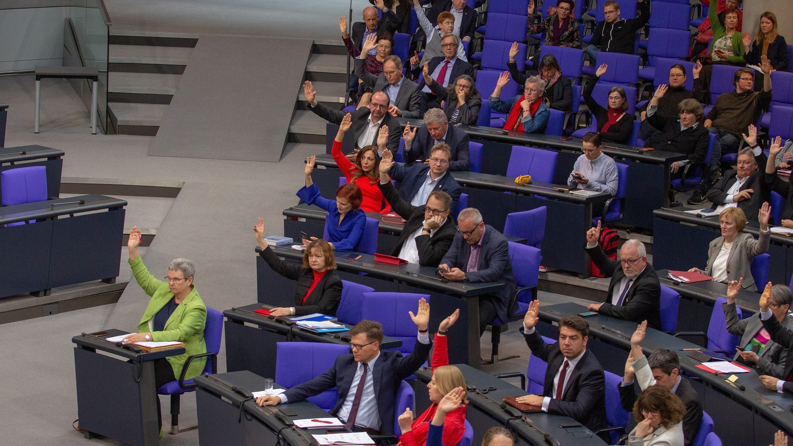Abgeordnete in den ansteigenden Rängen eines Plenarsaals, ein Teil hebt zur Abstimmung die Hand, einige Plätze bleiben leer