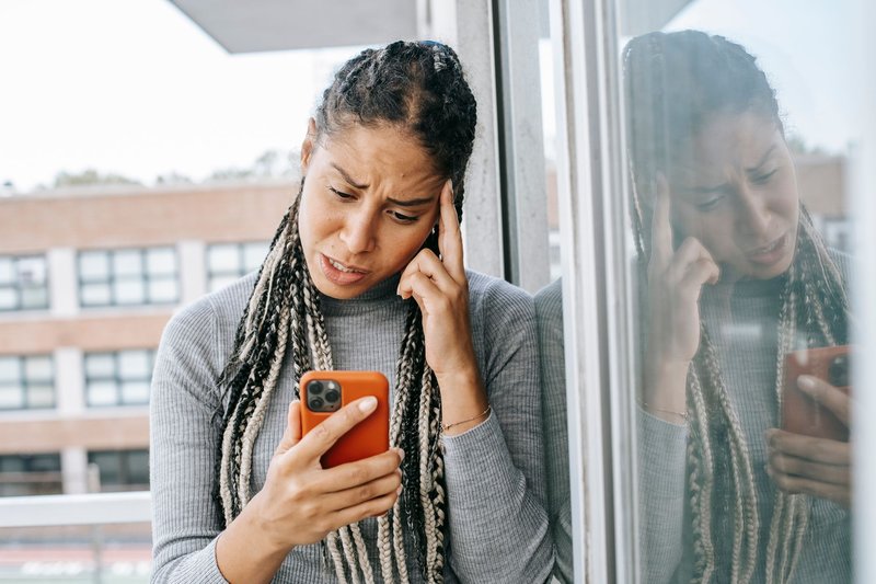 Junge Frau mit langen Zöpfen blickt auf einem Balkon bestürzt auf ihr Smartphone, eine Hand an der Schläfe