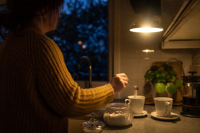 Frau gibt in einer dämmrigen Küche mit einem Löffel Zucker aus einer Zuckerdose in eine Kaffeetasse, daneben eine zweite Tasse