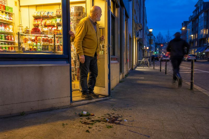 Kioskbesitzer in gelber Strickjacke steht am Abend in der Ladentür und blickt einer rennenden Person nach, davor eine zerbrochene Weinflasche auf dem Gehweg