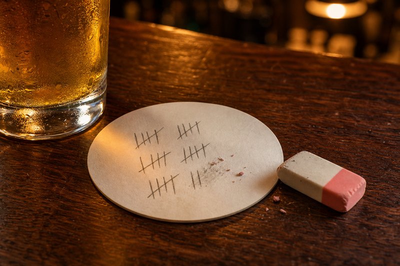 Bierdeckel mit Bleistiftstrichen auf einem Kneipentisch, zwei Striche halb wegradiert, daneben ein Radiergummi und ein Bierglas
